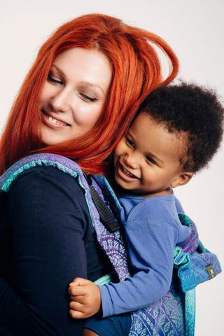 All about onbuhimos: Lenny Lamb Onbuhimo Peacock Tail Fantasy. A smiling white-presenting red-haired woman back carrying a happy, black-presenting toddler in an onbuhimo.