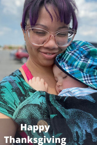 Black mother holds her white-presenting baby on her chest in a wrap strap meh dai. She is smiling, eyes closed. Baby is sleeping. Text reads, "Happy Thanksgiving"