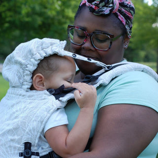 Black mother front carries her mixed race toddler in a Lenny Upgrade - Lotus Natural linen soft structured baby carrier. She is looking down at her sleepy child. The hood is up.