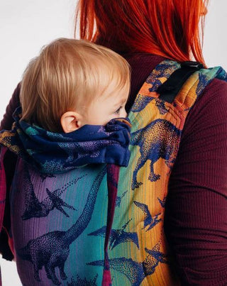 Red haired model is wearing a baby on her back in a Lenny Lamb buckle onbuhimo baby carrier in print Jurassic Park - New Era. The cotton baby carrier has realistic looking dinosaurs in a dark purple weft with background color gradient from gold to teal to purple to magenta