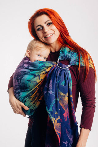 Model wearing baby in Lenny Lamb ring sling baby carrier in print Jurassic Park - New Era. Features Vibrant gradient background of purple to teal to yellow to red. Dark purple realistic-looking dinos and a on a dark purple weft. Leaning heads together and smiling