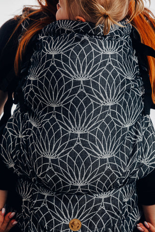 A model front carries a child in Lenny Lamb Preschool Carrier in Lotus- Black, a 100% linen carrier with black background and white outlined Lotus flowers