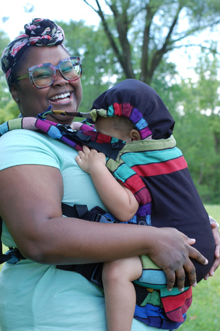 Smiling and laughing Black mother carrying her mixed race toddler on her front in Lenny Upgrade Carousel of Colors with mesh panel option. Hood is up.