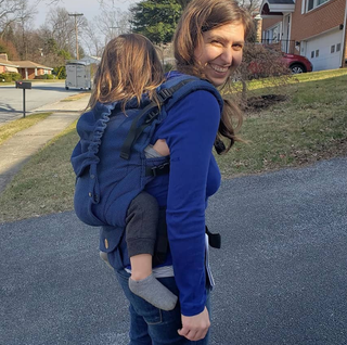 ﻿Mama & Roo's founder Alexandra smiles and wears her son on her back in the tester Preschool Carrier.