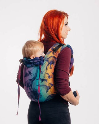 Red haired model is wearing a baby on her back in a Lenny Lamb buckle onbuhimo baby carrier in print Jurassic Park - New Era. The cotton baby carrier has realistic looking dinosaurs in a dark purple weft with background color gradient from gold to teal to purple to magenta