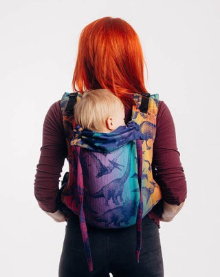 Red haired model is wearing a baby on her back in a LennyLamb buckle onbuhimo baby carrier in print Jurassic Park - New Era. The cotton baby carrier has realistic looking dinosaurs in a dark purple weft with background color gradient from gold to teal to purple to magenta
