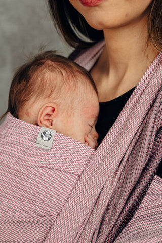Close up of model front wearing newborn (Inward facing) in long woven wrap. Print Little Herringbone Ombre Pink. Model is standing at a slight angle looking at the camera. 