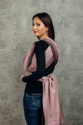Model front wearing newborn (Inward facing) in long woven wrap. Print Little Herringbone Ombre Pink. Model is standing at a slight angle looking at the camera over shoulder. 