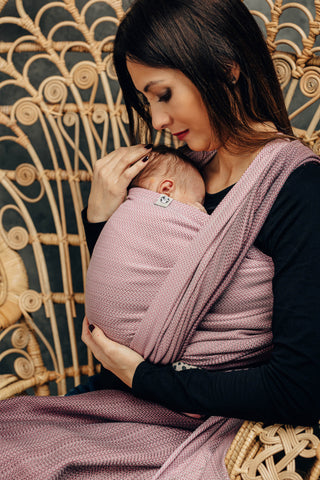 Close-up of model front wearing newborn (Inward facing) in long woven wrap. Print Little Herringbone Ombre Pink. Model is seated at a slight angle looking at newborn. 