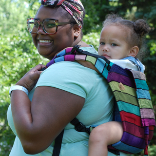 Carousel of Colors onbuhimo. A Black mother smiles as she carries her biracial child on her back in a rainbow striped waist-less carrier.