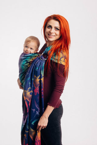 Model wearing baby in Lenny Lamb ring sling baby carrier in print Jurassic Park - New Era. Features Vibrant gradient background of purple to teal to yellow to red. Dark purple realistic-looking dinos and a on a dark purple weft. Side view