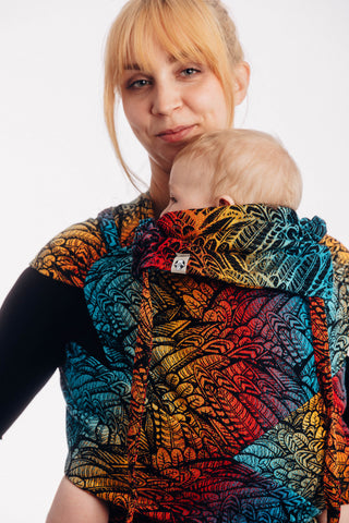 Woman carrying a baby in a Lenny Lamb meh dai / beh dai with wrap straps (wrap tai) in print Wild Soul Daedalus. The print has a black weft with feather pattern outlined in black on a background of electric blue, yellow, red, and orange.