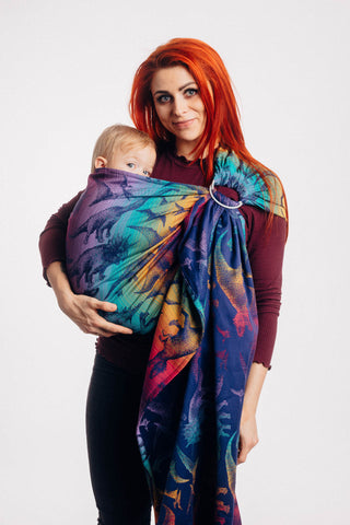 Model wearing baby in Lenny Lamb ring sling baby carrier in print Jurassic Park - New Era. Features Vibrant gradient background of purple to teal to yellow to red. Dark purple realistic-looking dinos and a on a dark purple weft. Standing
