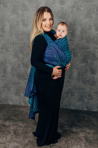 Mother smiles holding baby in Front Wrap Cross Carry in long woven wrap print Peacock's Tail Provance